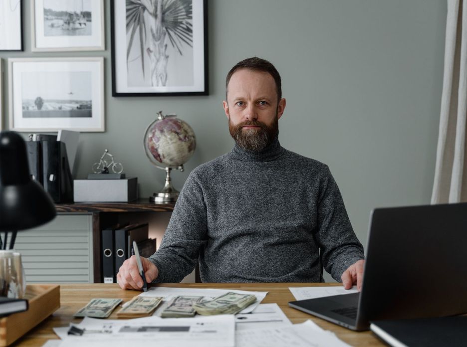 Bearded accountant in gray sweater working with banknotes and documents at office desk