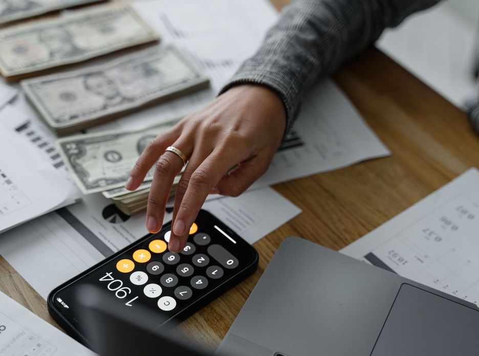 Close-up of a person using a smartphone calculator amid money and financial documents on a wooden table