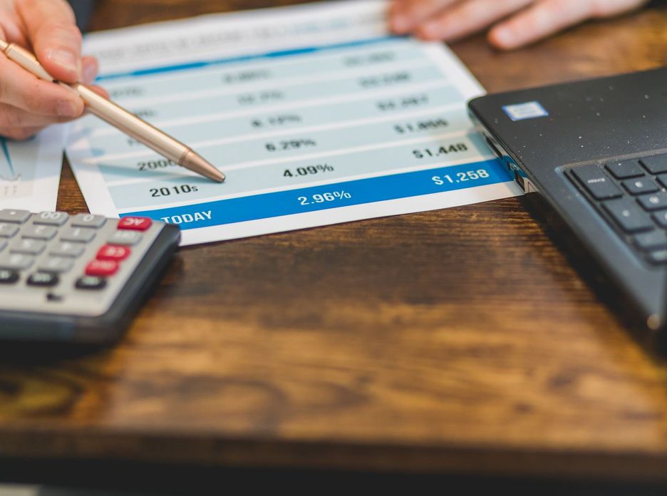 Close-up of a hand with pen analyzing financial rates on paper with a calculator and laptop nearby