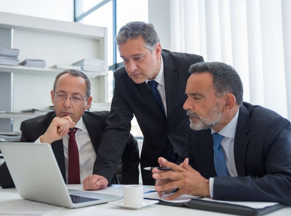 Three businessmen in suits collaborating in a modern office setting, focused on a laptop