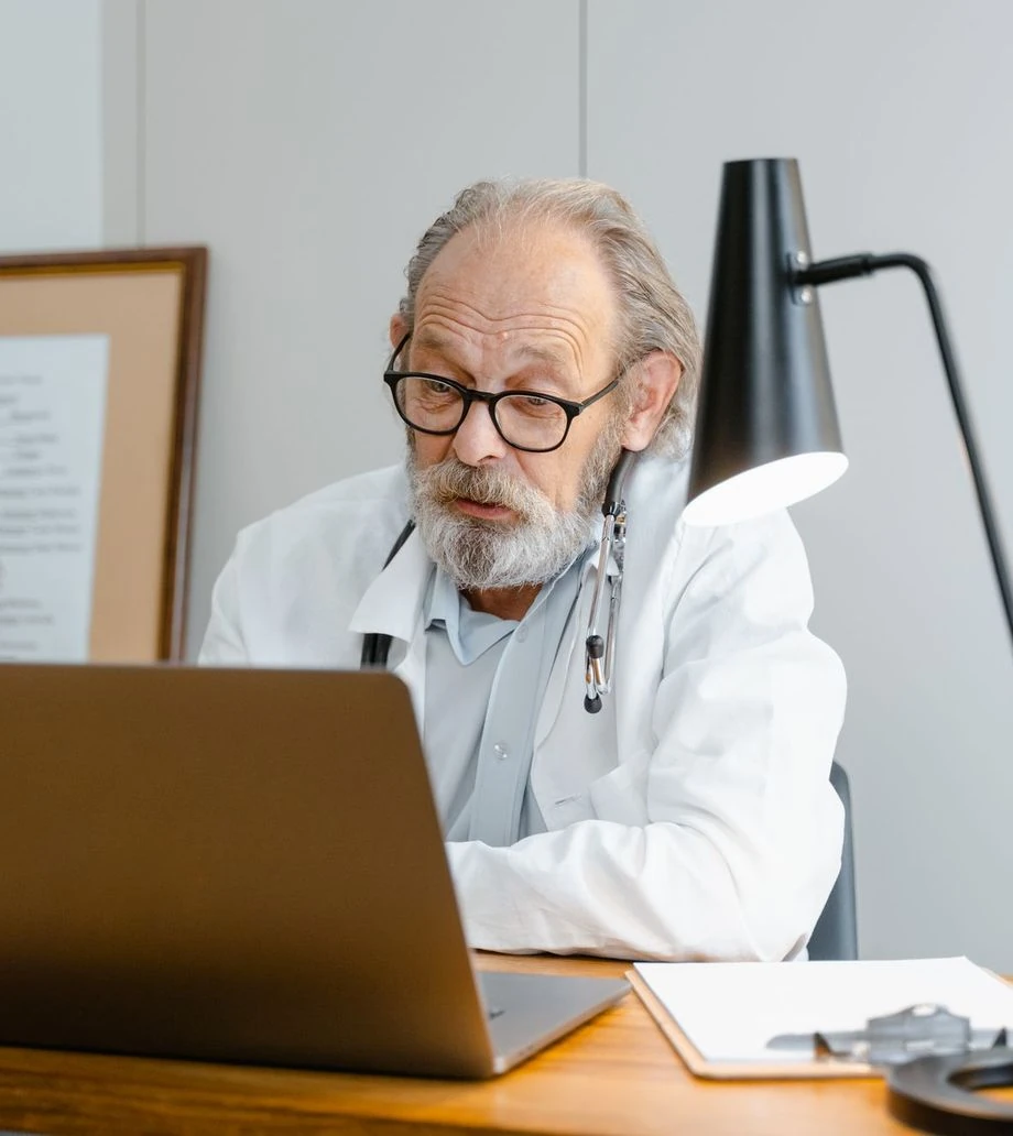 A doctor in a white coat working on a laptop in his office